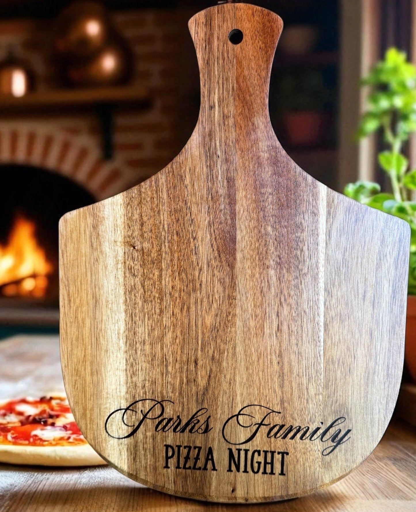 Wooden pizza peel with 'Parks Family Pizza Night' engraving on a kitchen counter with a pizza and fireplace in the background.